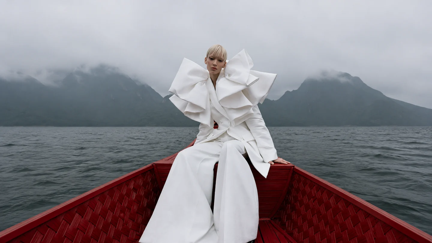Model in sculptural white tailoring seated in a red wooden boat on a misty mountain lake.