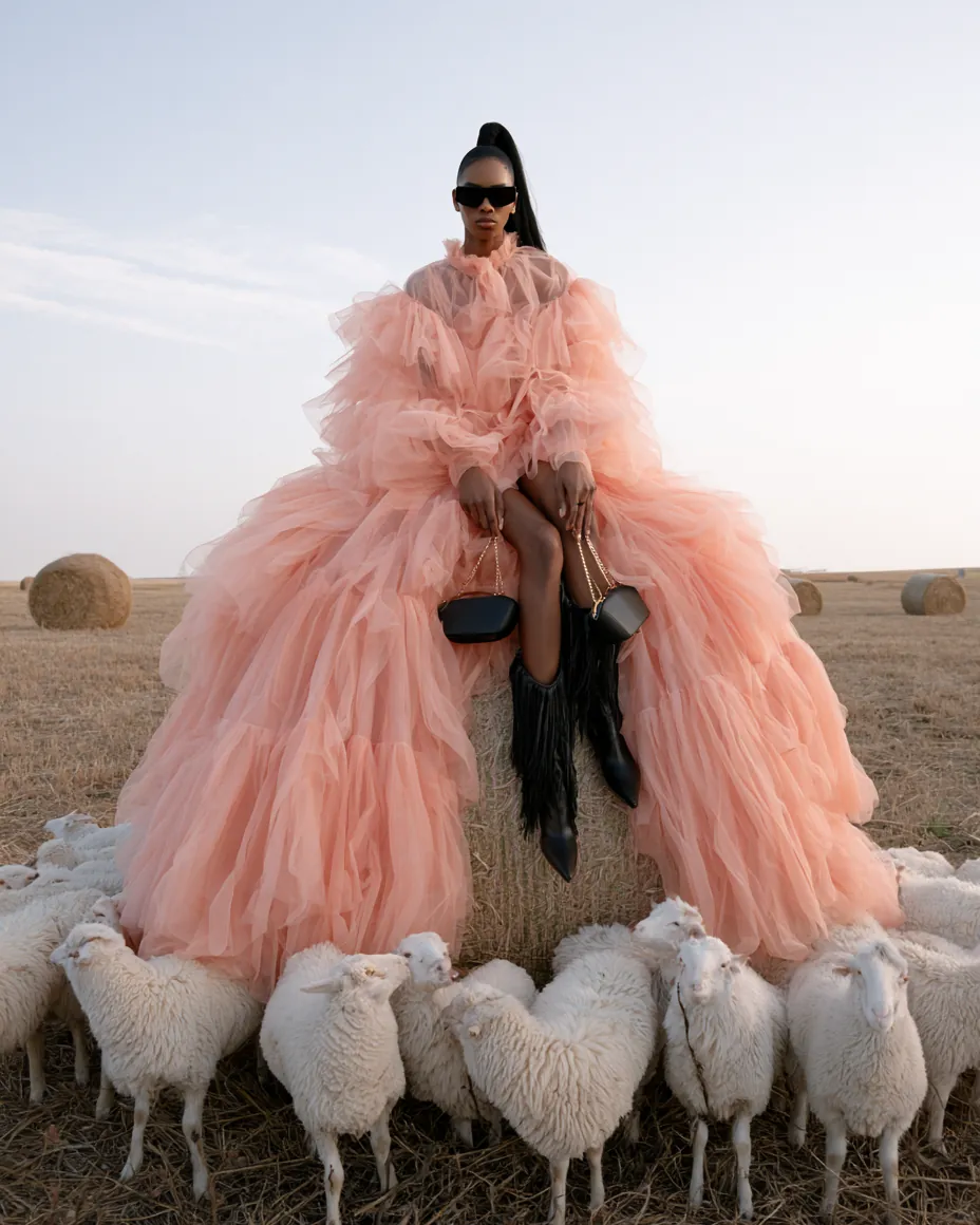 Fashion concept frame of a model in a voluminous pale gown standing in a cut field under natural daylight.